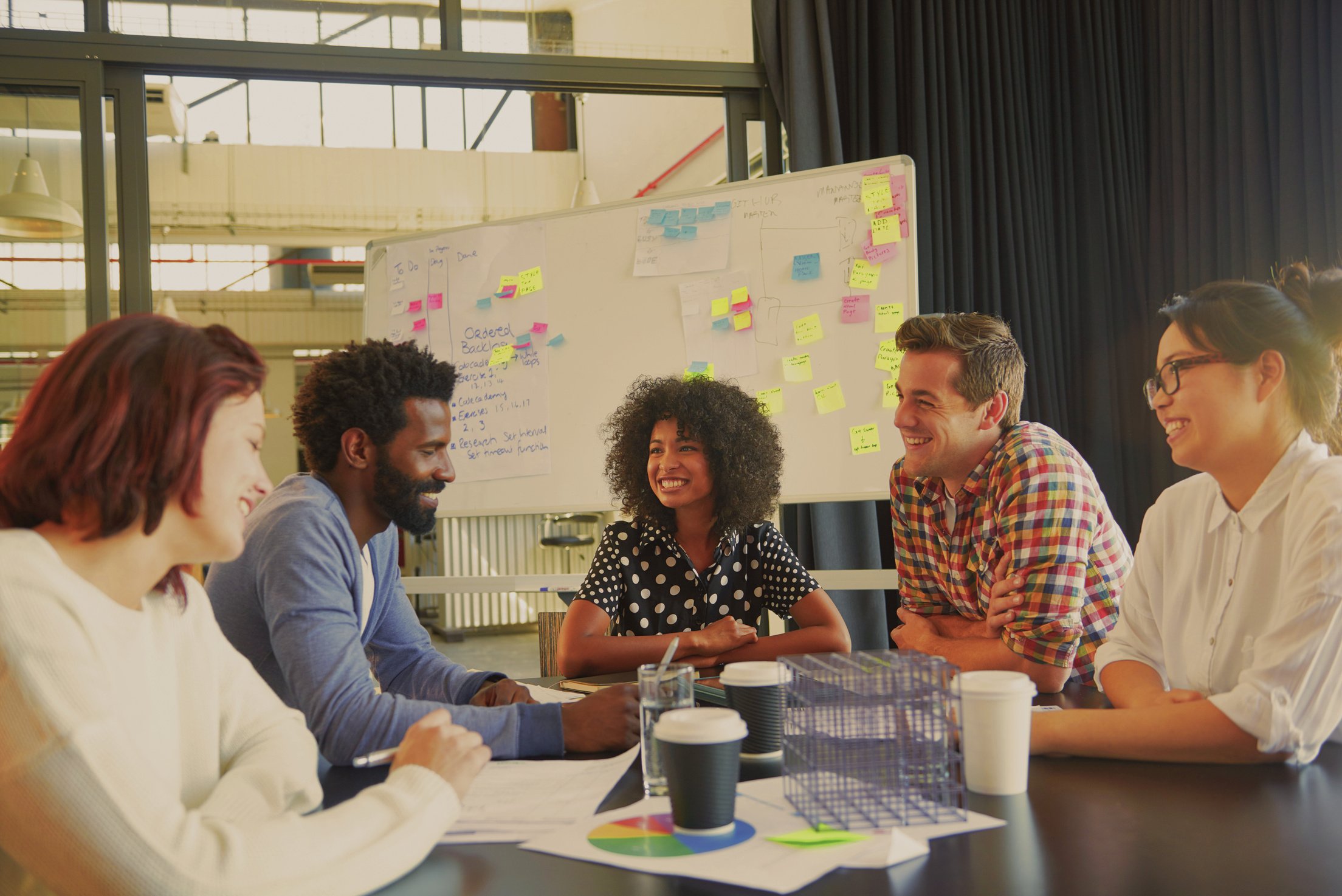 A group of co workers laughing and planning in a conference room with big picture windows and colorful post-it notes on a whitboard.